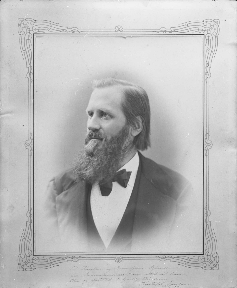 Portrettfotografi i halvprofil av yngre mann med halvlangt hår og langt fullskjegg, kledd i mørk jakke og vest, og hvitt skjorte med butterfly.