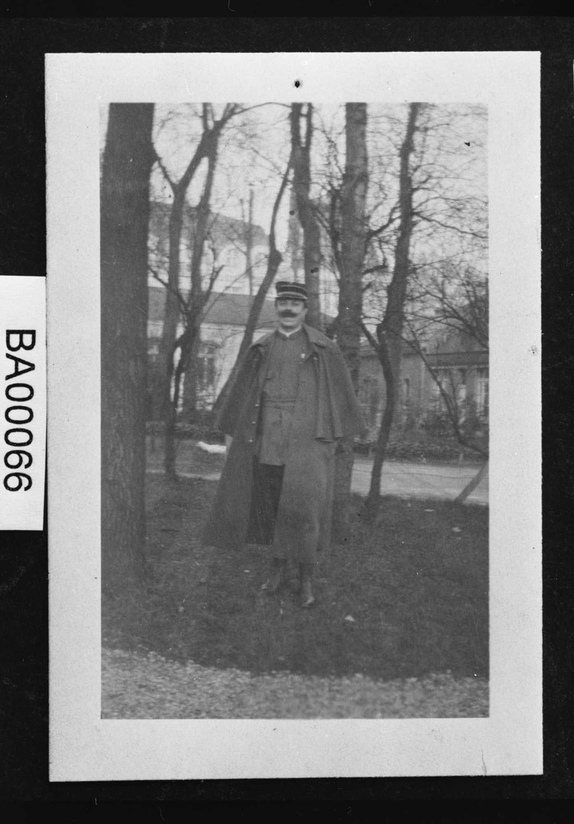 Fotografi; utendørsopptak: en voksen mann i militæruniform med stor lang kappe over skuldrene. I bakgrunnen skimtes en stor bygning og en del trær. 
