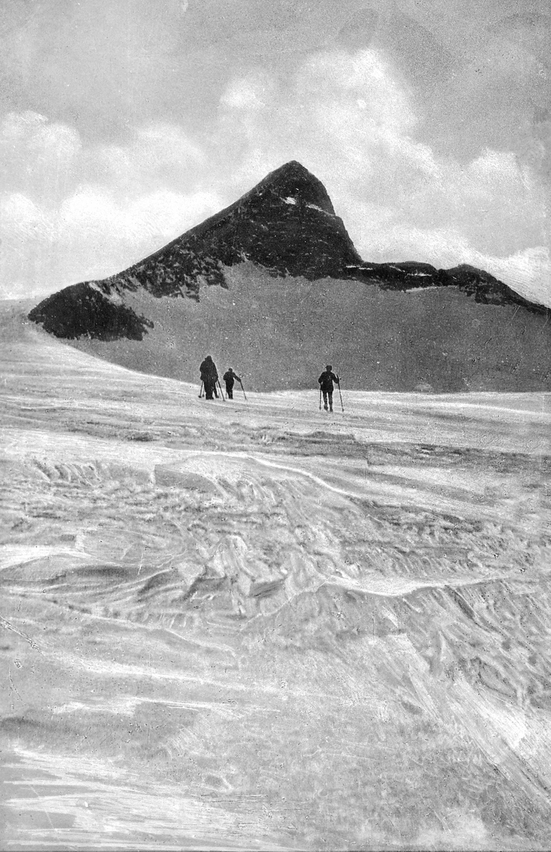Tre skiløpere, foran høy fjelltopp, vinter
