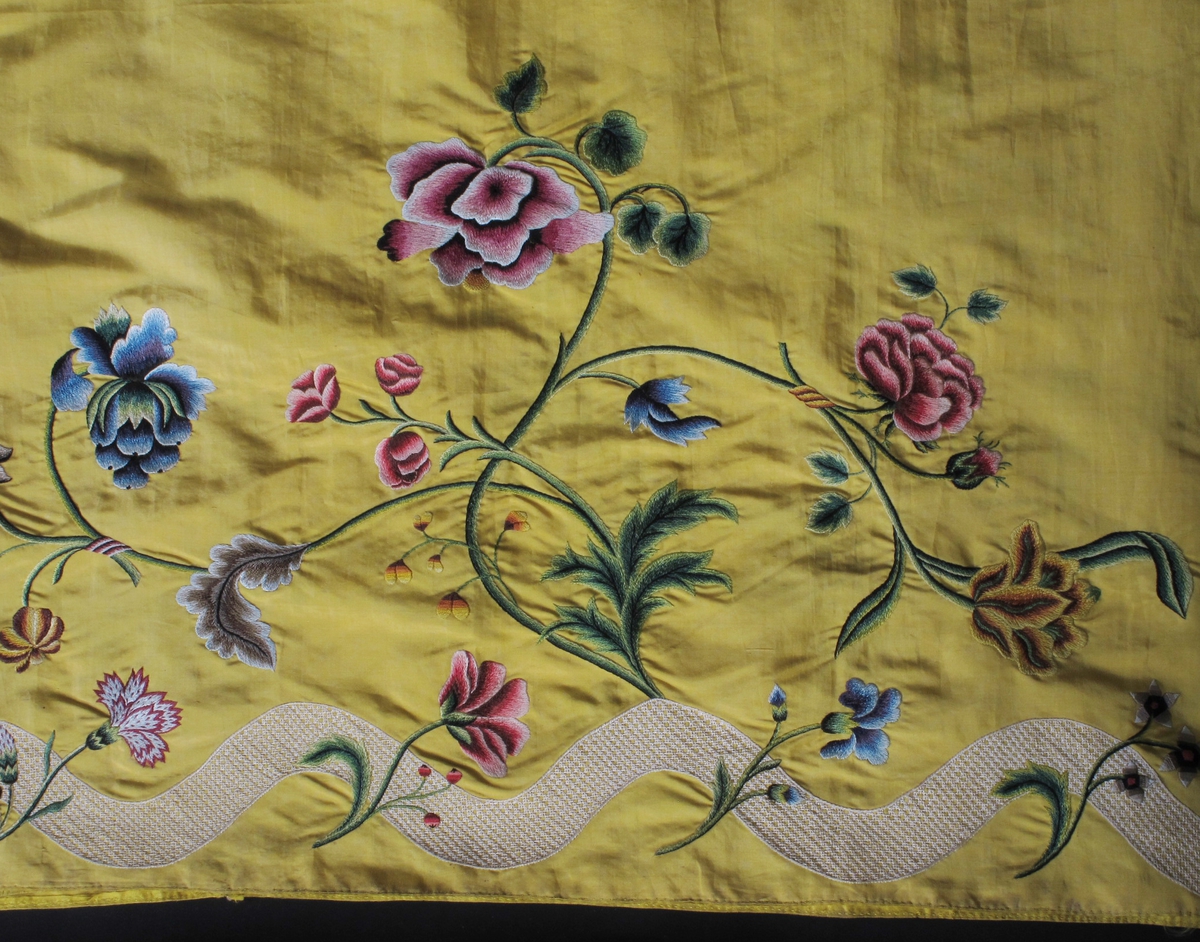 Del av en kjole,  ca. 1750   Silke,  sterkt gult. Brodert blomsterbord.   H. 88,5. Total L. 361. Opprinnelig et skjørt med bredt polykromt blomster  broderi langs nedre kant, slyngende gren. Under denn  en hvit bølgebord med smått rutemønster.  I senere tid, antag. 1900 tallet er skjørtet fjernet  fra linningen. det er påsydd en brunt bånd med ma  skin. Langs øvre kant på en av de fem breddene er  påtegnet med blyant et lignende blomstermotiv som  bede, men mer smått og tett. En opprinnelig lerrets  lomme henger på.  Tilstand a meget god.