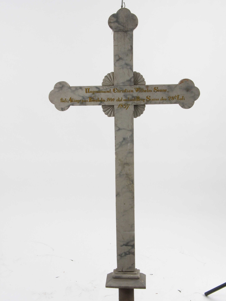 Kors i tre, malt lyst grått med marmorering og innskrift. 
