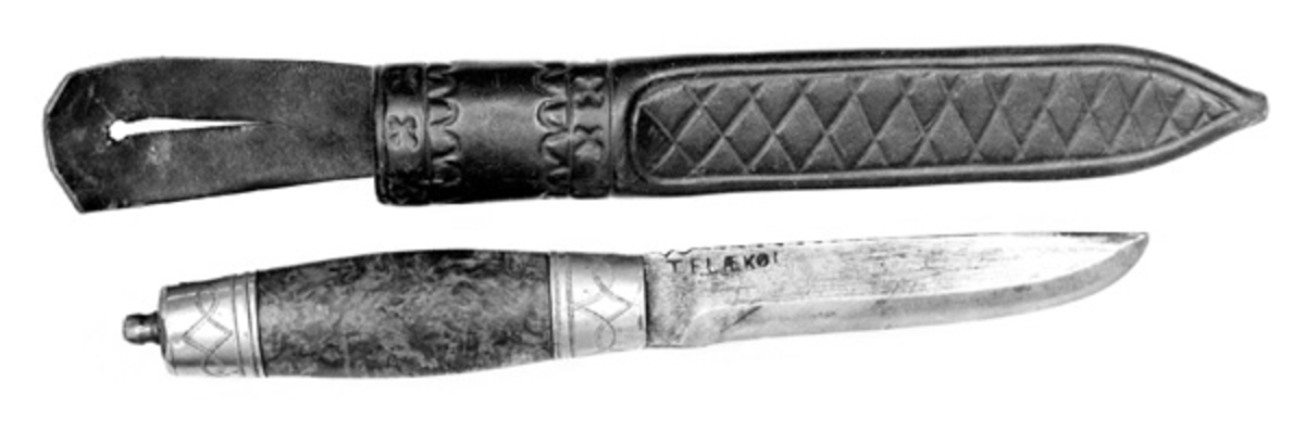Kniven har brunbeiset valbjørkskaft, nysølv holker og messing- plate og knapp på toppen av skaftet. Svart lærslire.