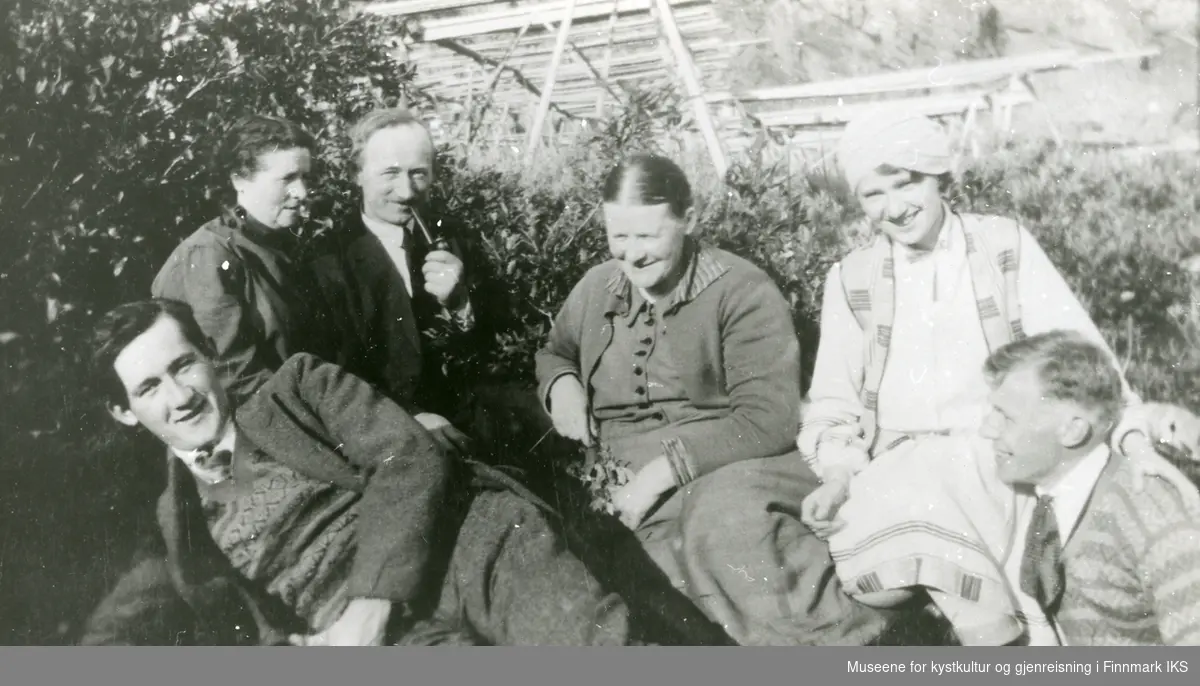 6 personer sitter ute på en gressbakke, fiskehjeller i bakgrunnen. Ca 1929.