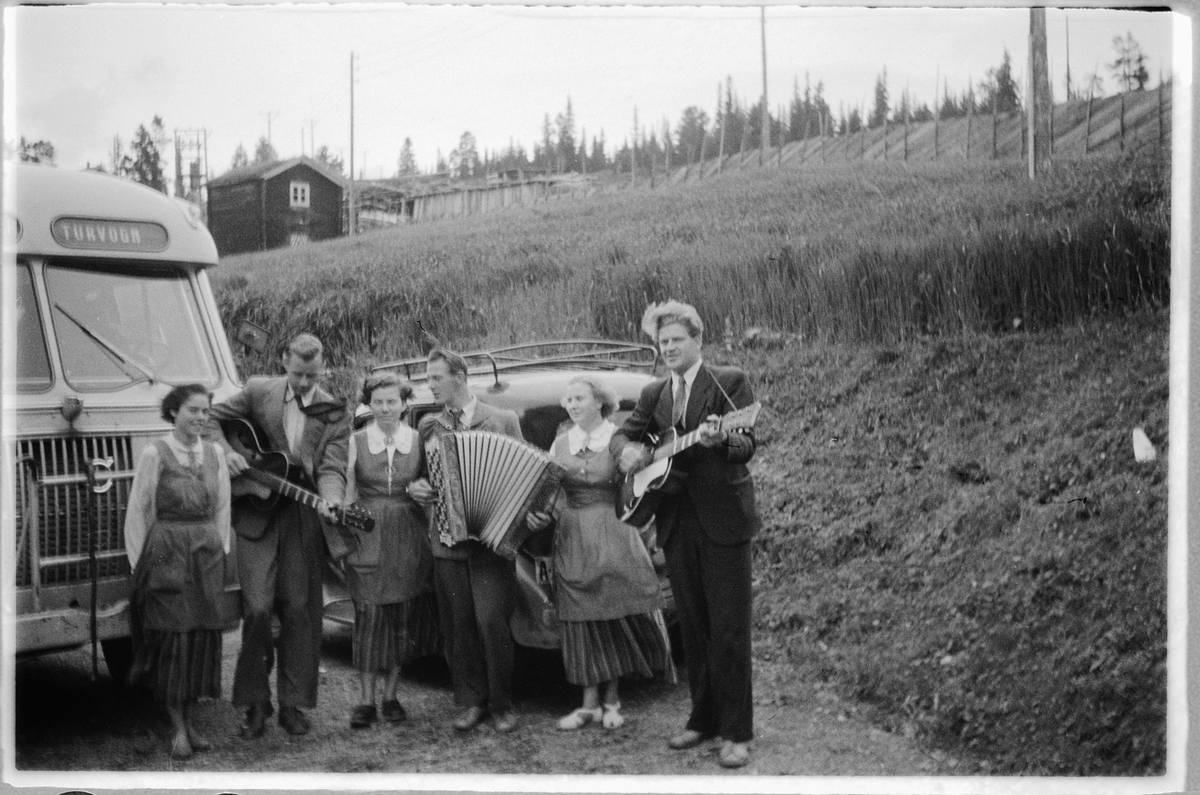 Tre menn spiller gitar og trekkspill, mens tre kvinner lytter ved en buss med skilt Lillehammer turvogn. T. h. Ulf Sæhli på gitar. Se HHB-7409-28. 