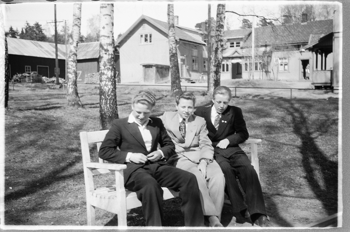 Moelv sentrum, Tre unge menn, i dress sitter på en benk. I midten Reidar Larsen (Baashus), de to andre er ukjente. Kan alle tre være lærlilnger for Frisør Johansen? I bakgrunnen Fruseth kafeen.