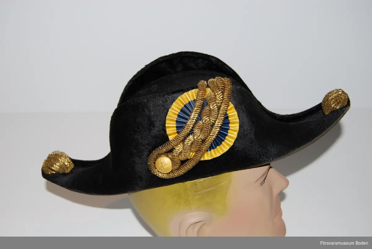 Svart kläde, knapp och kokard med agraff på höger sida. I hattens spetsar "tassar" av gulddragararbete.
