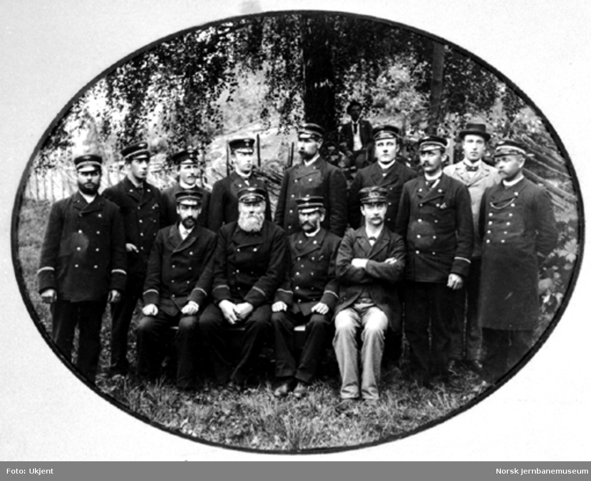 Gruppebilde tatt i landlige omgivelser av 12 jernbanefolk samt to i sivil, sannsynligvig på Hamar ca. 1895