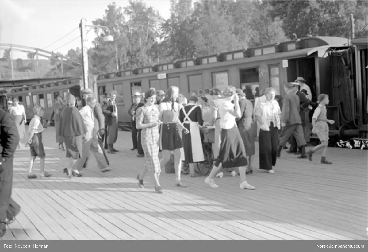 Eidsvoll stasjon med mange reisende foran et fjerntog; servering på plattformen