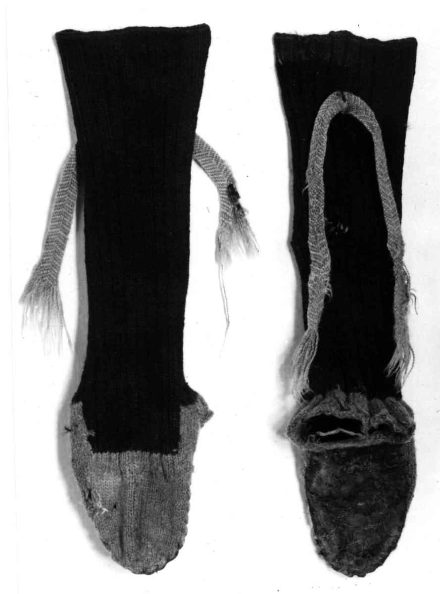 Svarte raggsokker i ull med såler av lær. Fot av grått ullgern, uten hæl.
Sokkebånd flettet av rødt og gult ullgarn, fiskebeinmønster.