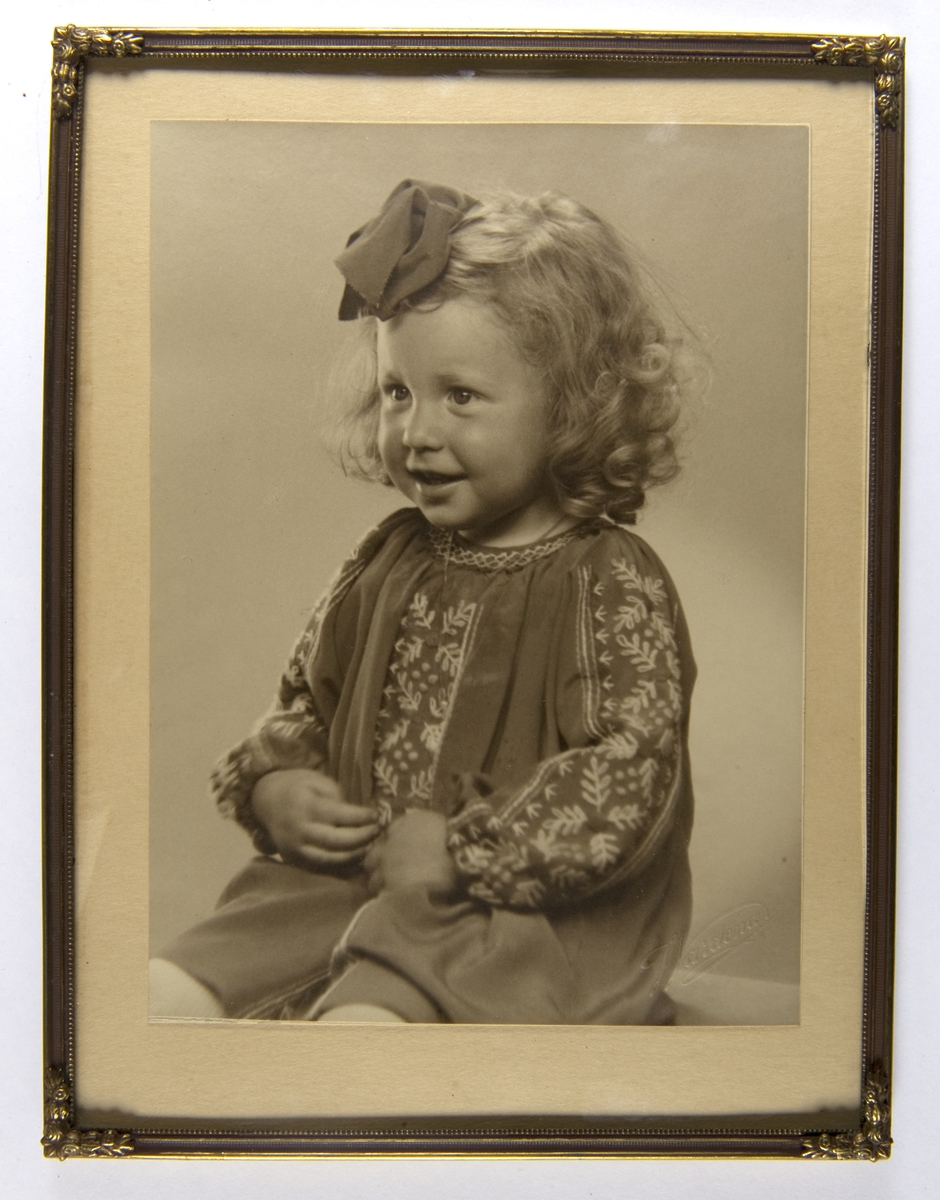 Sittende liten jente i strikkekjole, sløyfe i håret.
