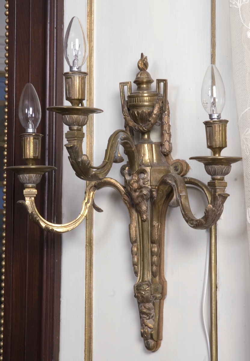 Veggkandelaber; bronse, forgylt, trearmet, dekorert med blad, bukkehode og en urne øverst.
