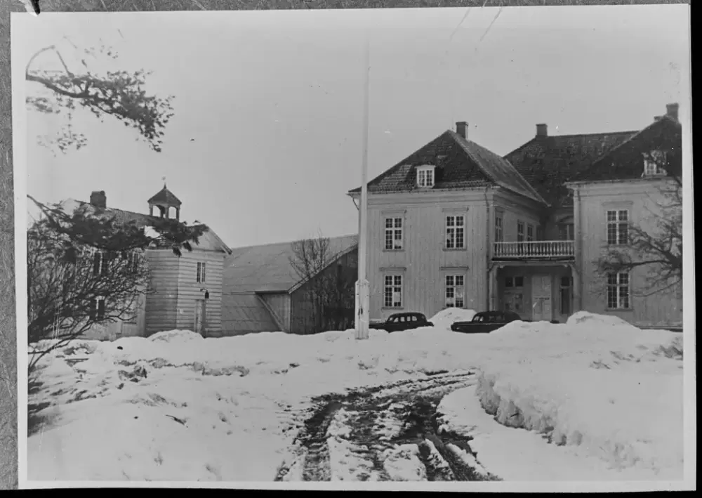 Svart-hvitt bilde av Eidsvollsbygningen vinterstid, omgitt av snø. Eidsvollsbygningen står sentralt i bildet, med en flaggstang uten flagg foran. Ujevn snø og hjulspor leder opp mot hovedinngangen, hvor noen biler er parkert.