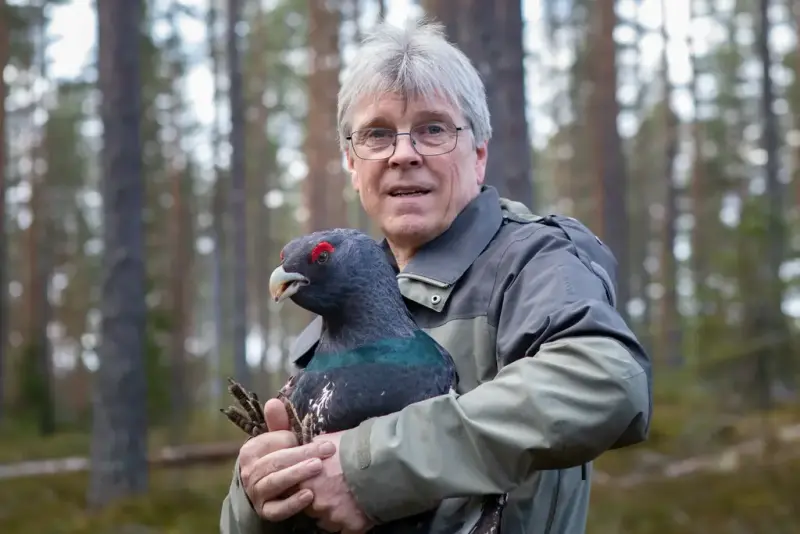 Portrett av Trond Berg. Han har kort grått hår og briller. Han har på seg en grønn jakke. I hånden holder han en tiur. 