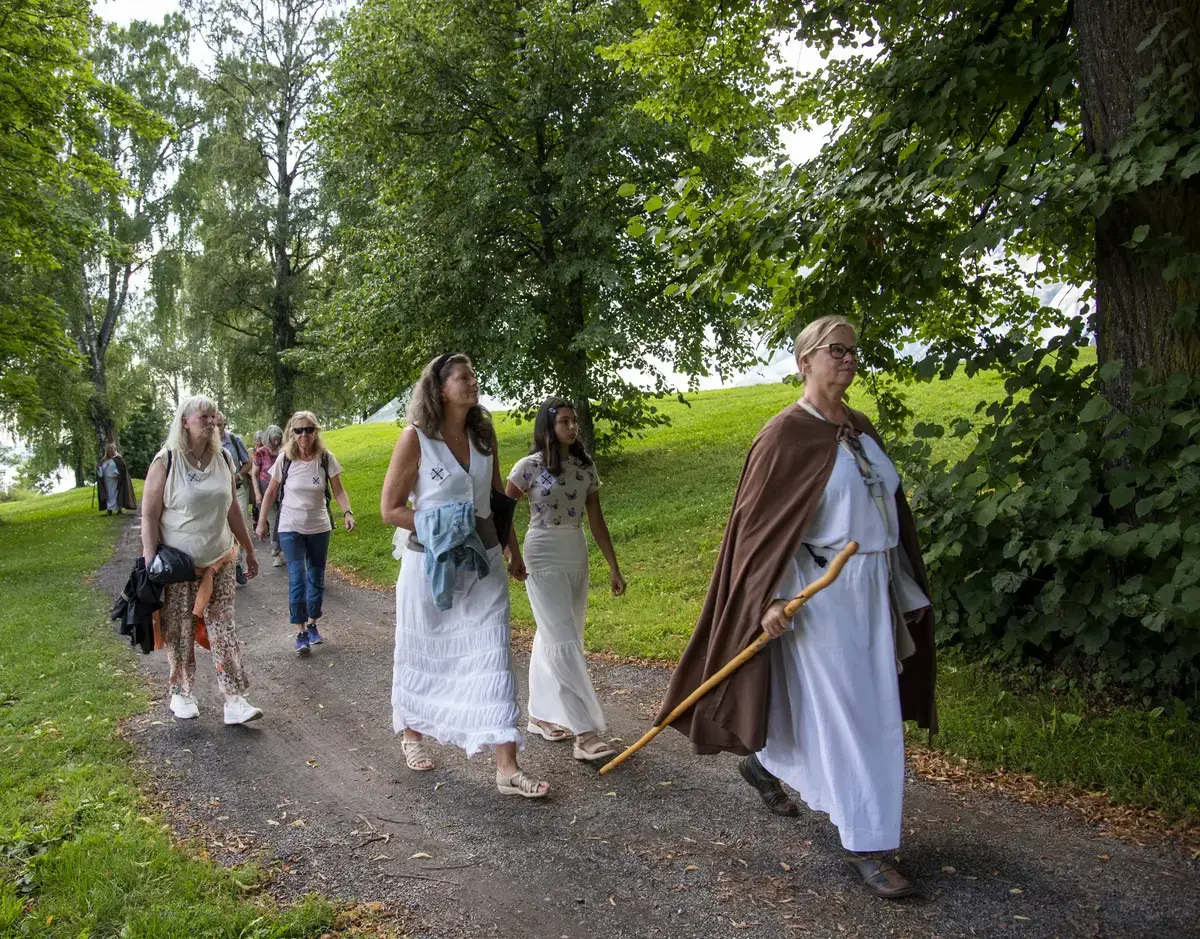 Kvinne i hvit middelalderkjortel og brun kappe, med pilegrimsstav i hånda, leder an en gruppe sommerkledde mennesker på vandring langs en grusveg.