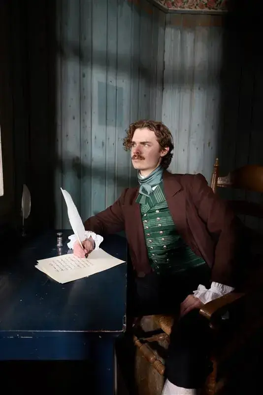 Man in costume sits at a writing desk holding a quill pen.