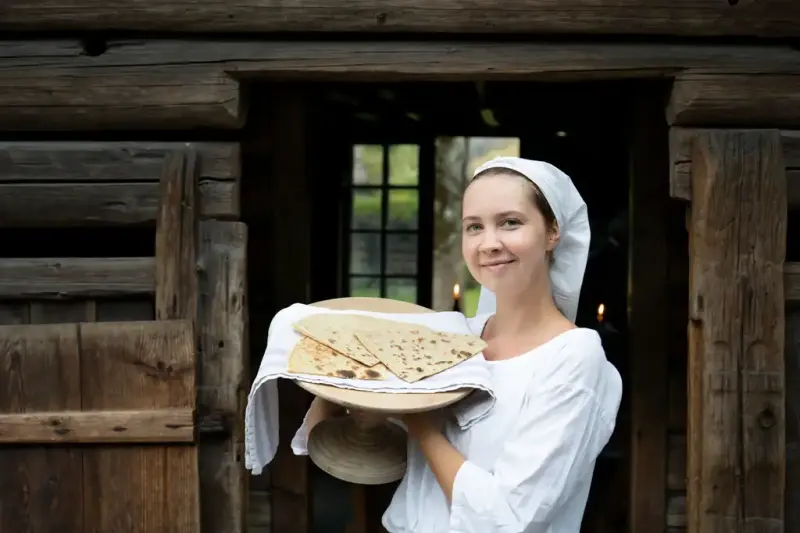 Woman in historical costume holding a plate of freshly baked lefse outside a log building in Numedal at Norsk Folkemuseum.