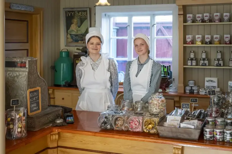 To jenter i gammeldagse kostymer står bak en kjøpmannsdisk med godteri
