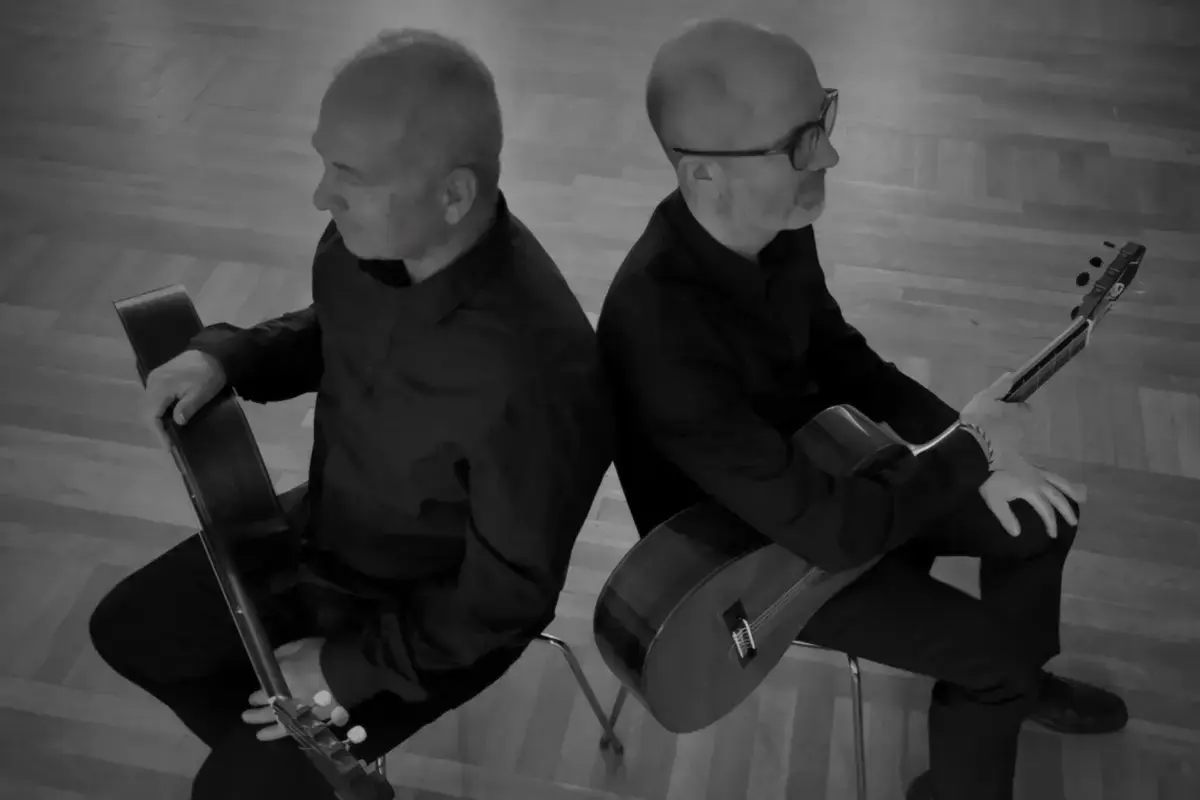Svart-hvitt foto av to menn som sitter med ryggen mot hverandre. De har hver sin gitar i fanget.